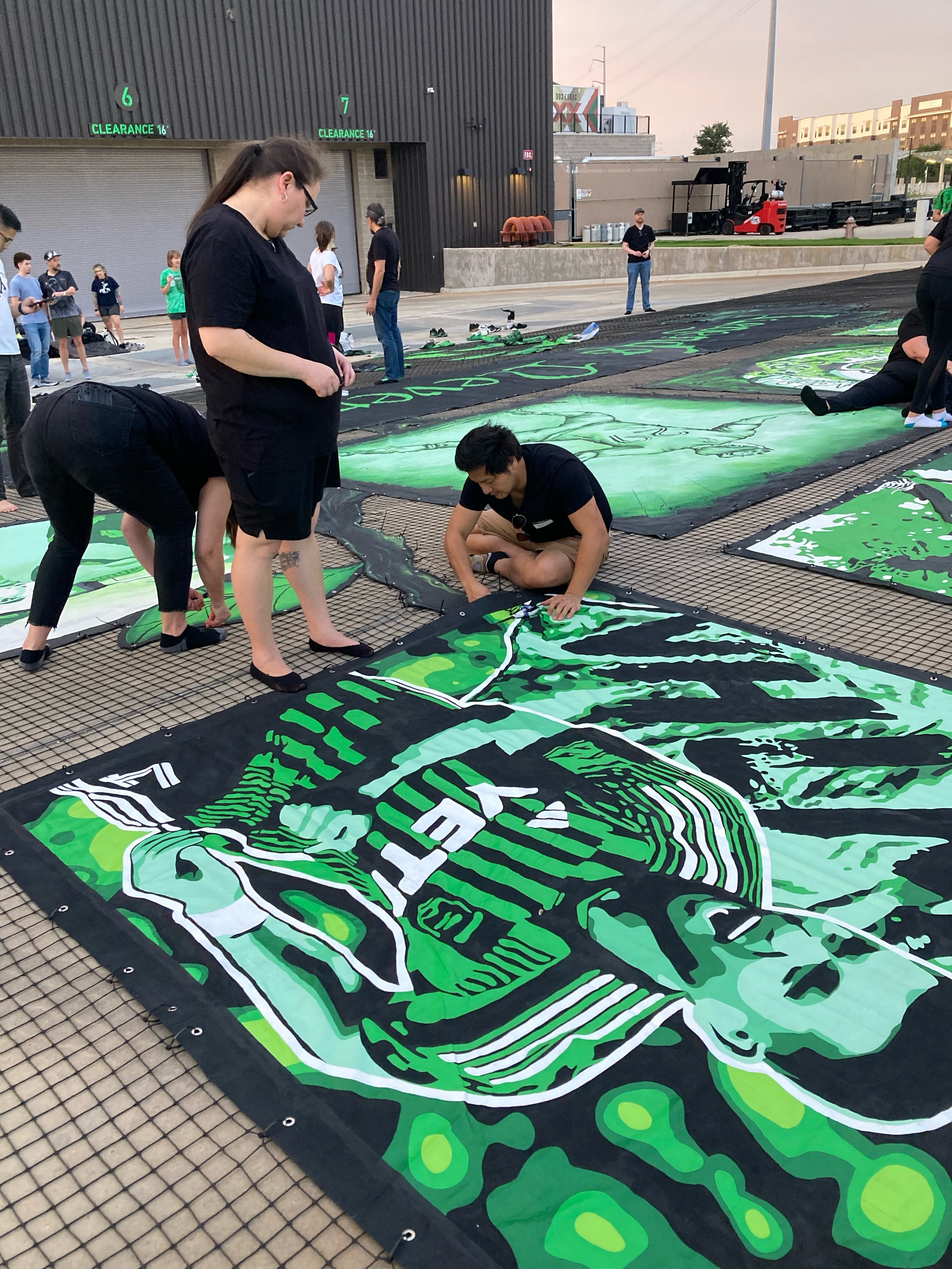 Volunteers in black t-shirts kneeling and painting large green and black tifo banners laid out on the ground.