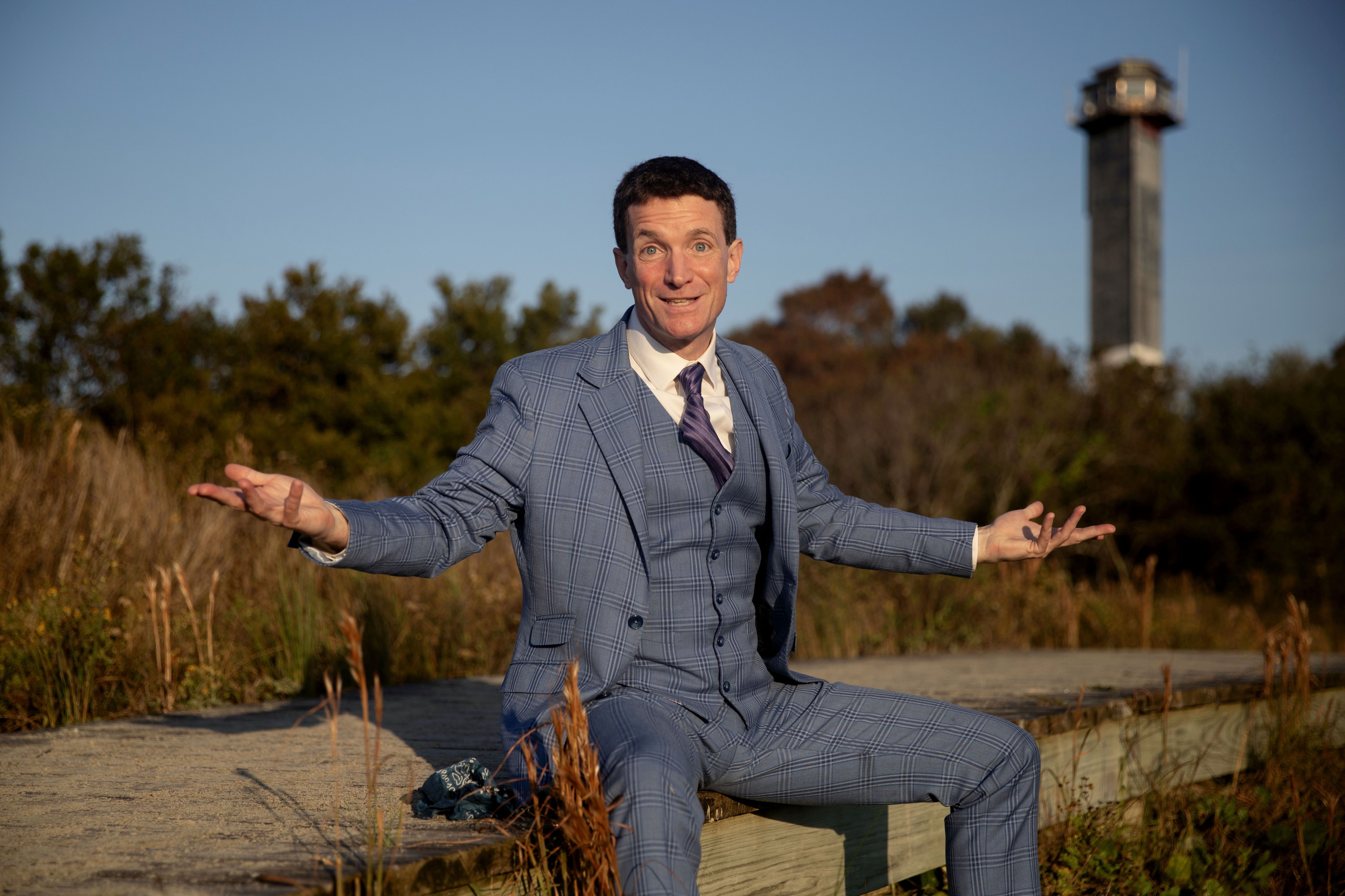 Grady Hendrix sitting on a wooden walkway with arms outstretched, smiling, in a blue plaid suit with a lighthouse in the background.